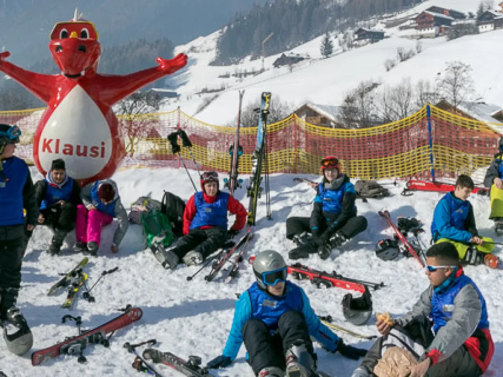 Jugendliche sitzen im Schnee neben einer Maskottchenfigur.